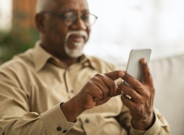 Senior African Man Using Smartphone Texting Sitting On Sofa Indoor